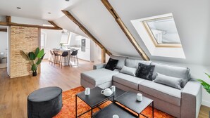 Un loft moderno con techo inclinado, un sofá seccional gris, un reposapiés negro y una cocina al fondo.