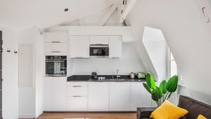Une cuisine moderne avec des armoires blanches, un four encastré et un four à micro-ondes. Le salon est doté d’un canapé marron foncé avec des coussins jaunes et d’une grande plante verte.