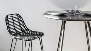 Une horloge murale ronde avec un cadre doré, une chaise en osier noire et une table avec deux verres et des assiettes.