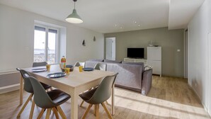 Ein modernes Wohnzimmer mit Essbereich, grauen Sofas, einem Flachbildfernseher und einem Balkon, der durch das Fenster sichtbar ist.