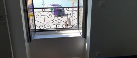 Une chambre dotée d’un balcon donnant sur un port, comprenant un lit, une fenêtre avec vue sur les bateaux et un phare, ainsi qu’une rambarde en métal.