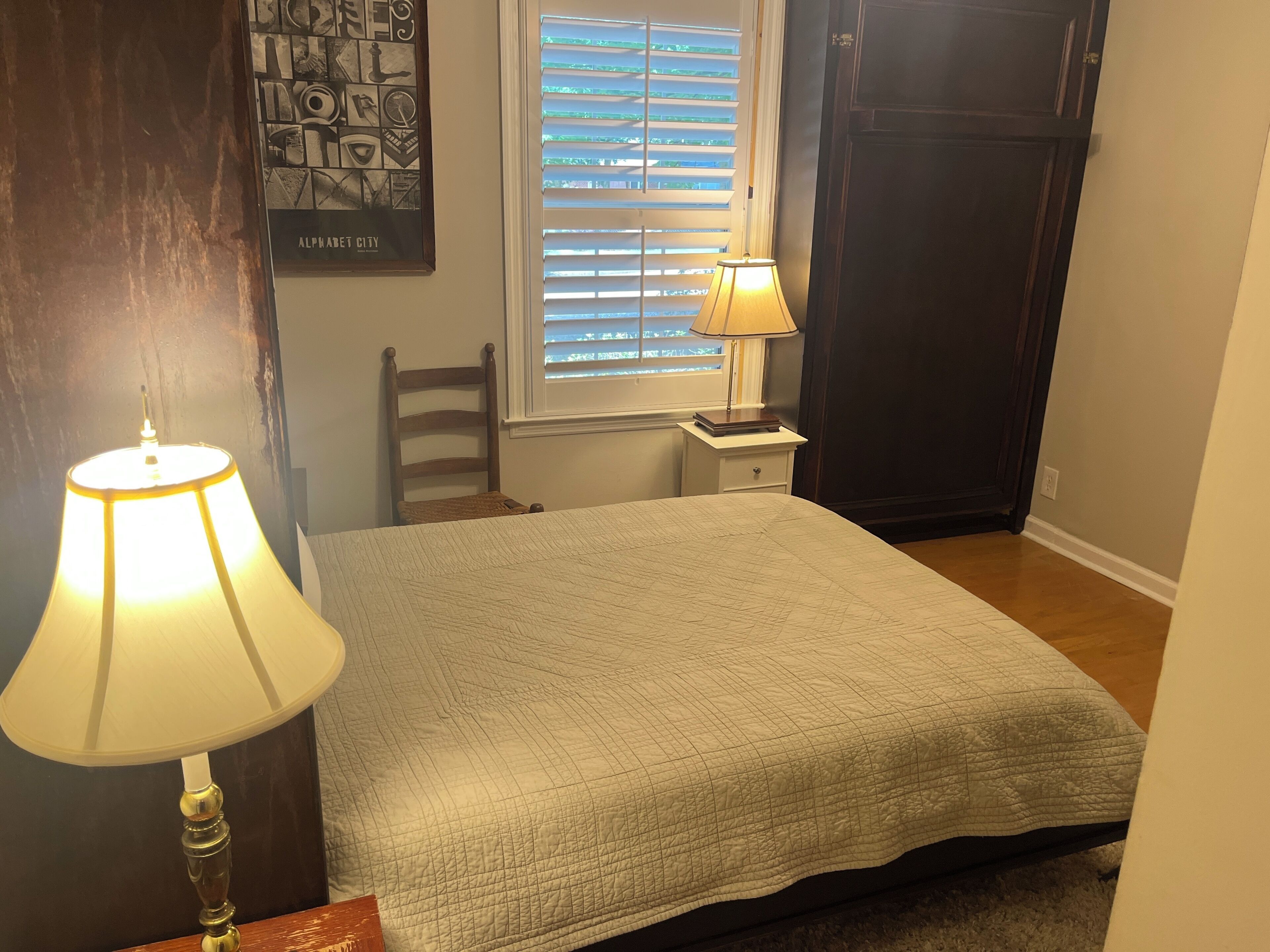 Une chambre à coucher avec un lit, deux lampes de chevet, une fenêtre avec des stores, une chaise, une petite table et une porte.