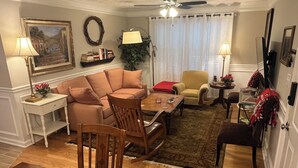 A living room with a sofa, armchairs, a coffee table, and a dining table. There are lamps, a plant, and a bookshelf with books.