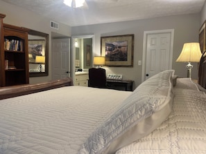 A bedroom with a large bed, a wooden headboard, a desk with a lamp, a mirror, and a bookshelf.