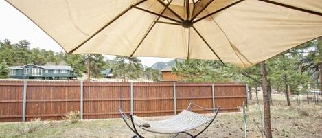 A hammock under a large canopy in a backyard with a wooden fence and trees in the background.