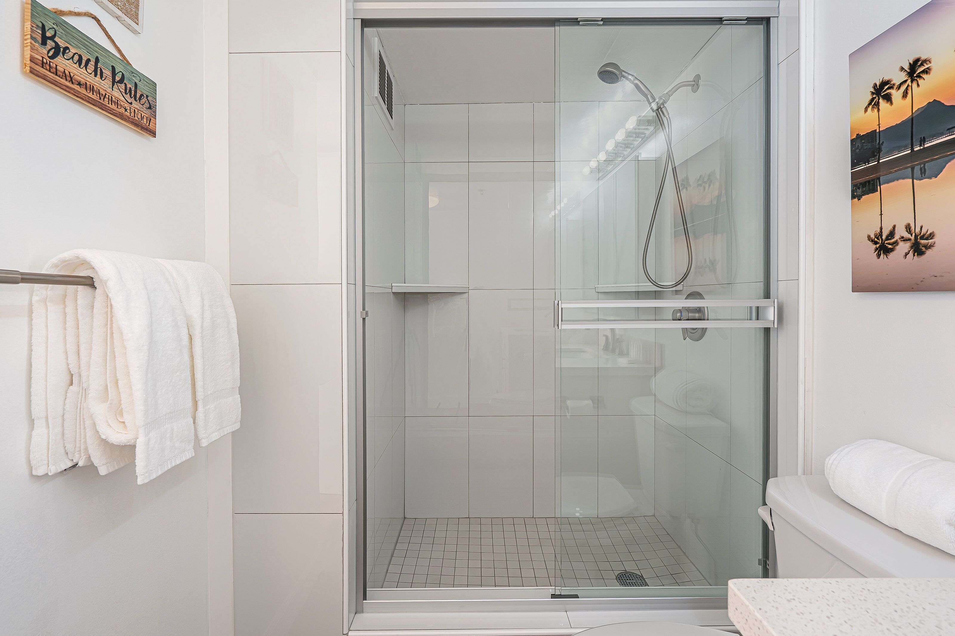 A glass-enclosed shower area with a handheld showerhead, a towel rack with white towels, and a framed picture of palm trees on the wall.