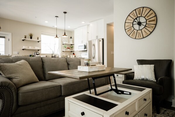 A modern living room with a sectional sofa, a coffee table, a side table, and a large wall clock. - Farmhouse at Lincoln Suites — Smart Home, King Bed, Sheridan WY (Sheridan)