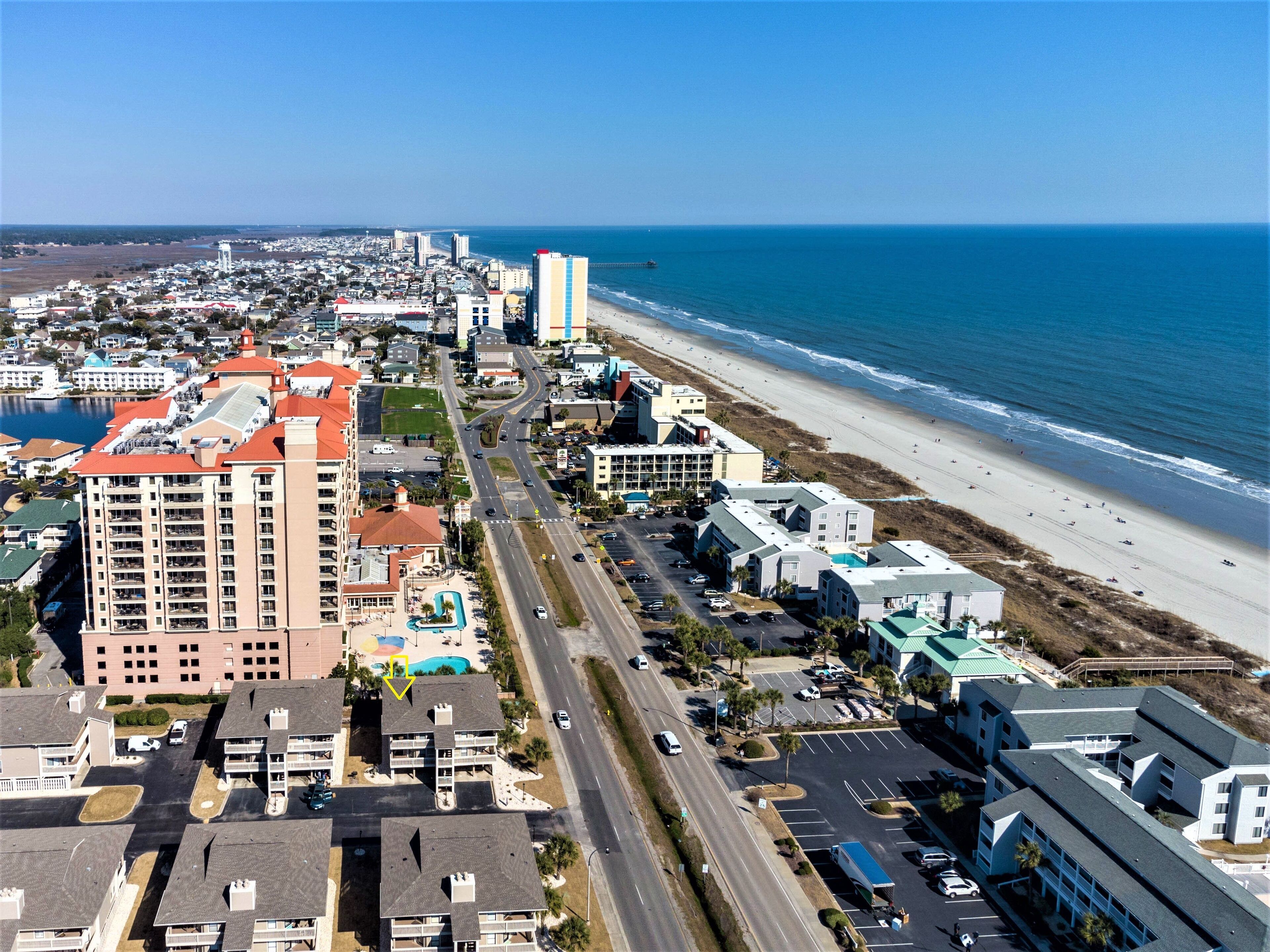 An aerial view of a coastal city with a beachfront, including a hotel with multiple floors and a swimming pool.