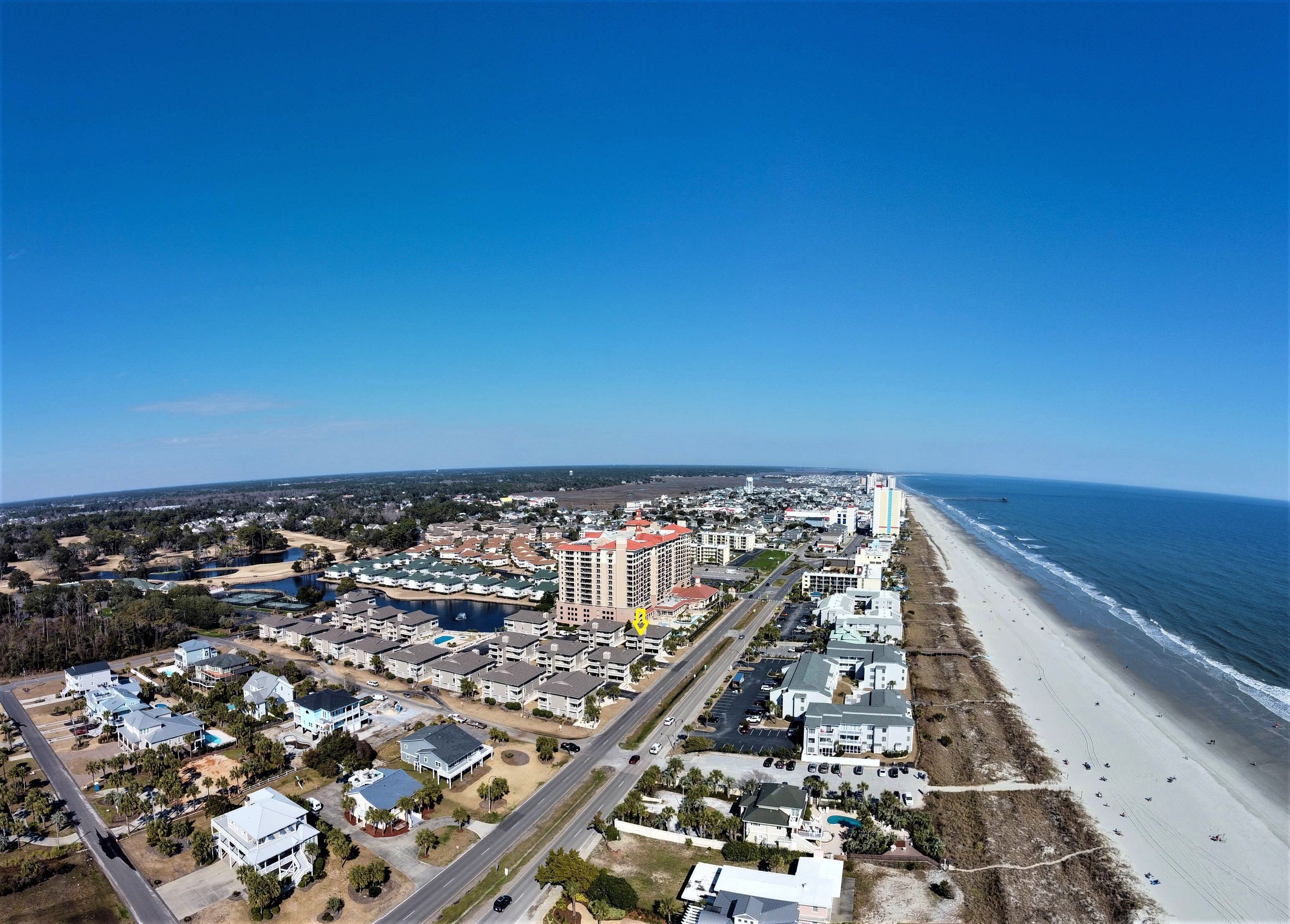 An aerial view of a coastal town with a beachfront, residential buildings, and a large hotel with multiple floors.