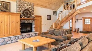 A spacious living room with a stone fireplace, wooden furniture, and a staircase leading to an upper level. - Fischer's Paradise (Presque Isle)