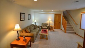 Una sala de estar con un sofá, dos sillones, una mesa de centro y una lámpara. Se ve una escalera con barandillas de madera que conduce a otro nivel.