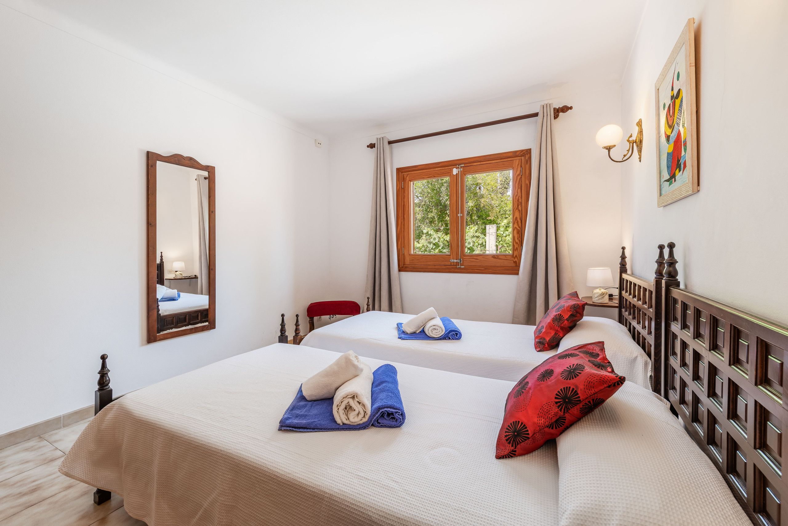 A hotel room with two beds, a window, a mirror, and a painting.
