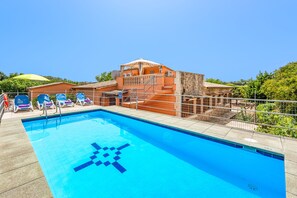 Una piscina con un diseño geométrico azul, rodeada de sillas de descanso y un edificio con tejado de baldosas.