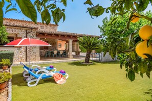 Un patio con mobiliario exterior, una mesa cubierta y vistas a la piscina de un edificio con paredes de piedra y techo de tejas.
