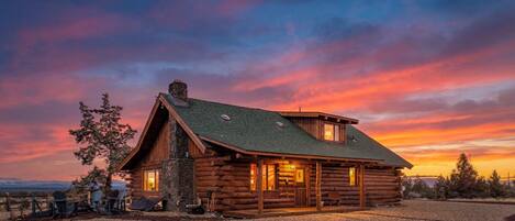 A log cabin with a stone foundation, a green roof, and a porch with chairs.