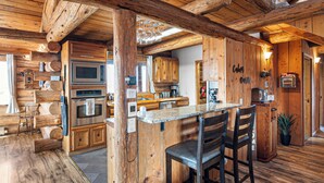 A rustic cabin interior with wooden walls, a kitchen area with modern appliances, and a bar with two stools.
