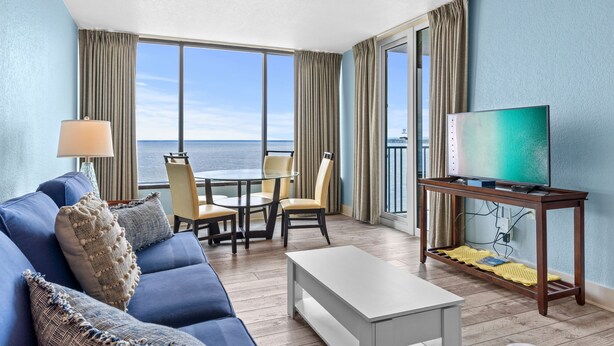 A modern living room with a sofa, coffee table, TV, and a balcony with a view of the ocean. - Stylish 1BR Condo w/ Bay View! Balcony, Pool, Tiki Bar, Tram to Beach & Wharf (Miramar Beach)