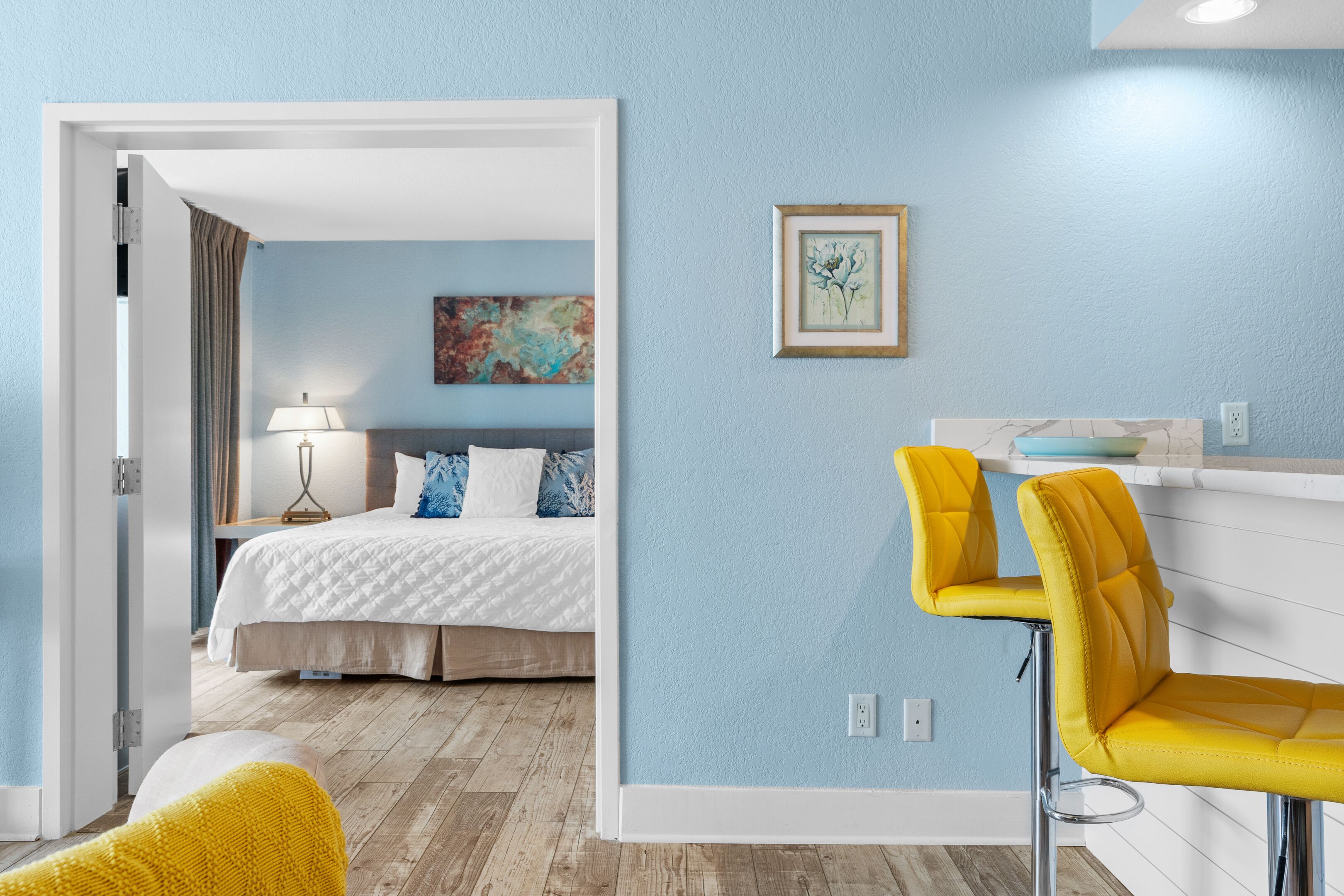 A modern bedroom with a bed, bedside table, and a nightstand. There is a window with curtains, a wall-mounted artwork, and a small table with a lamp.