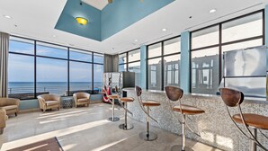 A spacious lounge area with large windows, a bar with stools, and a view of the sea. - Stylish 1BR Condo w/ Bay View! Balcony, Pool, Tiki Bar, Tram to Beach & Wharf (Miramar Beach)