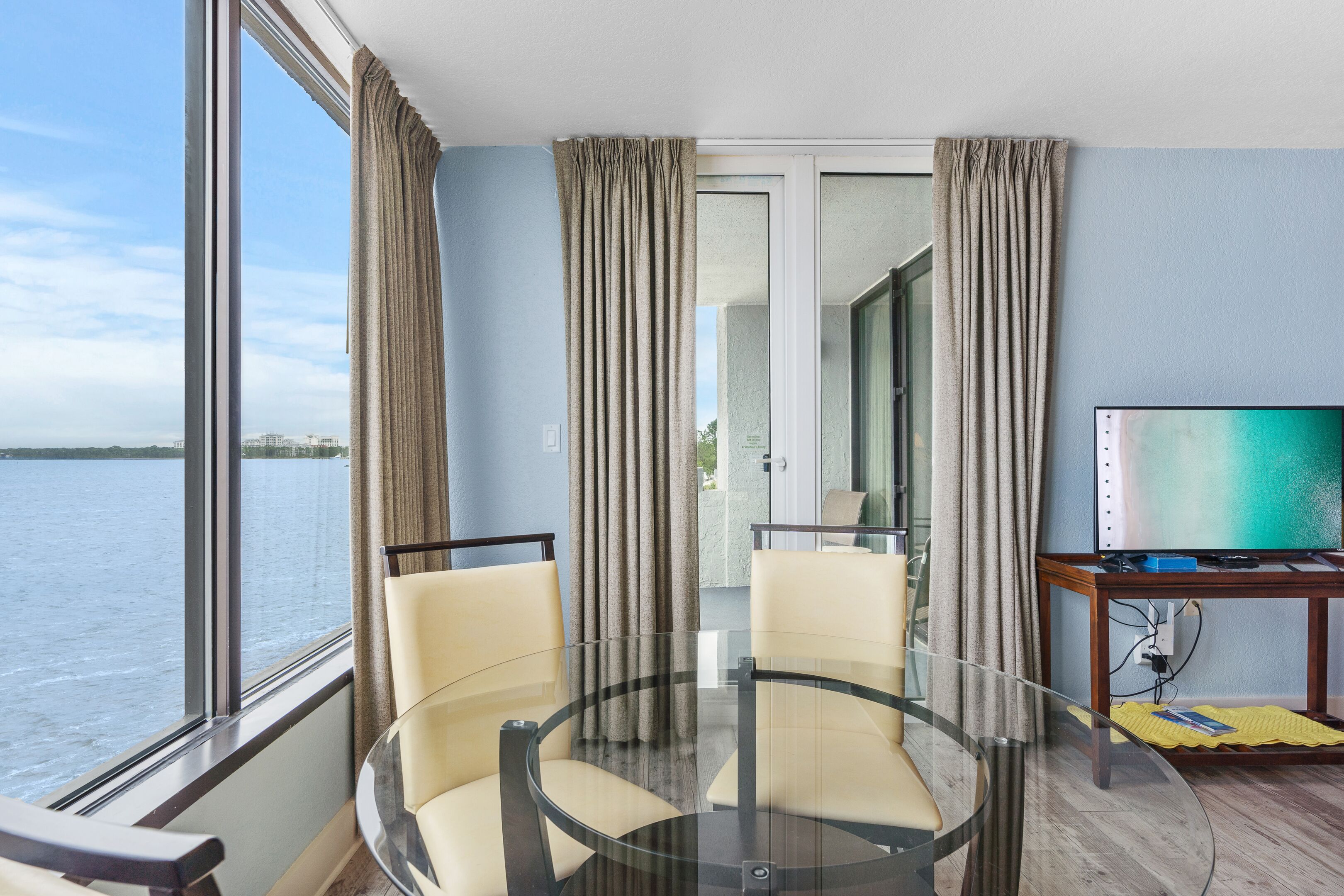 Una habitación con una mesa de vidrio, dos sillas, un televisor y vista al agua.