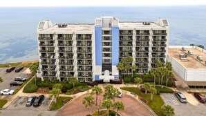 A multi-story hotel with a central blue and white vertical stripe, surrounded by parking and palm trees, overlooking a body of water. - Stylish 1BR Condo w/ Bay View! Balcony, Pool, Tiki Bar, Tram to Beach & Wharf (Miramar Beach)
