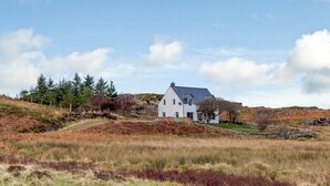 Une maison blanche avec un toit gris, entourée d’un paysage vallonné où la végétation est clairsemée et où l’on trouve quelques arbres.