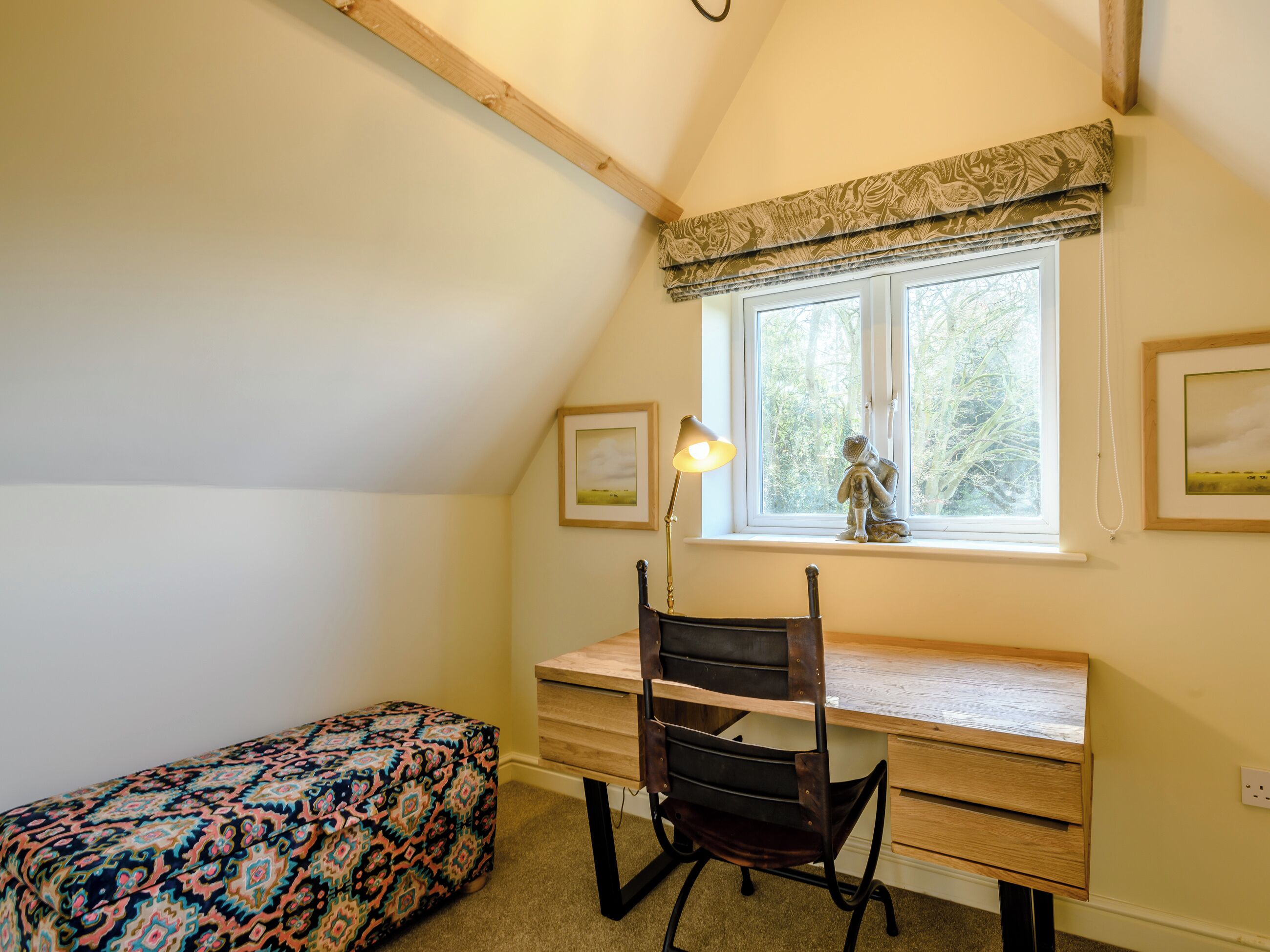 Una camera da letto con una scrivania in legno, una sedia, un letto con copriletto decorato, una finestra vista alberi e una statua sul davanzale.