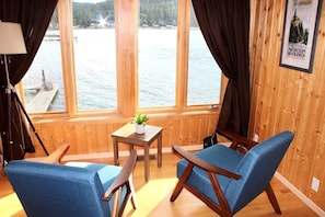 Een kamer met houten wanden en grote ramen met uitzicht op een wateroppervlak. Er staan twee blauwe stoelen en een klein tafeltje met een plantje.
