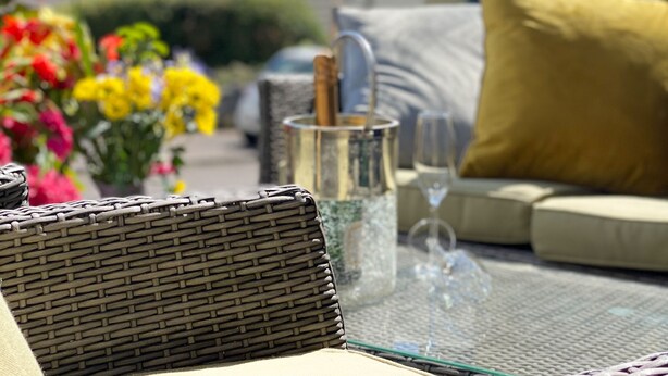 A wicker chair, a glass table with a metal container, a wine glass, and a yellow pillow.
