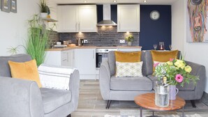 A modern kitchen with white cabinets, a stove, and a sink. There is a dining area with two grey sofas and a coffee table.
