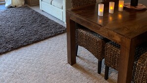 Ein gemütliches Wohnzimmer mit Esstisch aus Holz, Korbstühlen, einer Couch und einem großen Fenster mit Vorhängen.