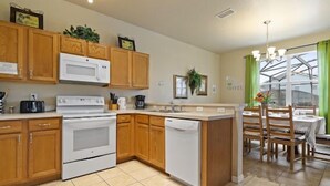 A modern kitchen with wooden cabinets, a white stove, and a dishwasher. There is a dining area with a table and chairs, and a large window with green curtains. - Blue Jay Villa - Vacation Home Near Disney (Davenport)