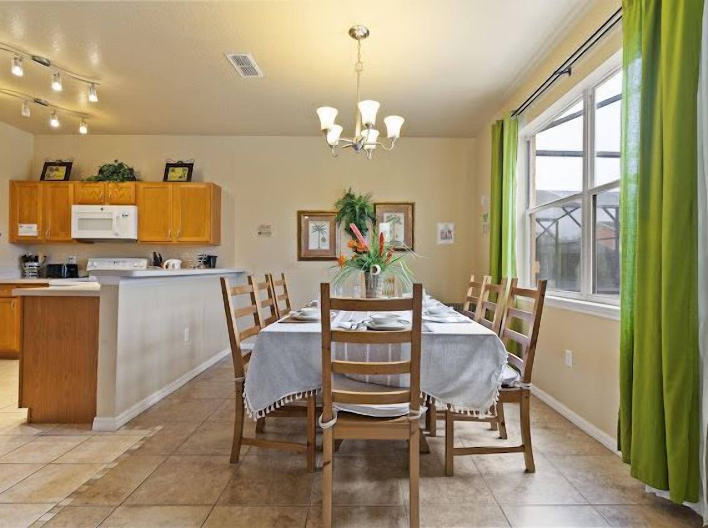 Un coin repas avec une table dressée pour quatre personnes, des chaises en bois, un lustre, une cuisine avec des meubles en bois et une fenêtre ornée de rideaux verts.
