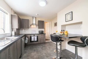 A modern kitchen with dark cabinetry, a built-in oven, a stainless steel range hood, and a bar area with stools. - Thy Grace 3 Bedroom 2 Shower Room Central Newland (Kingston upon Hull)
