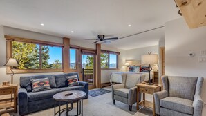 A modern living room with a sofa, armchairs, a coffee table, and a ceiling fan. Large windows provide a view of trees and mountains. - 🏔️Ski Access~Hot Tub~Studio Premier 106🏔️ (Snowmass Village)