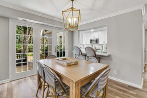 A dining area with a wooden table and wicker chairs, a kitchen with white cabinets, and a view through glass doors to a garden.