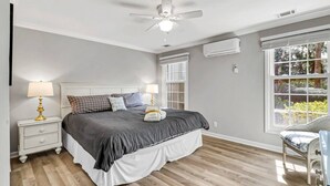 Un dormitorio con cama, mesitas de noche, ventilador de techo, unidad de aire acondicionado y ventana con persianas.