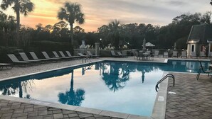 Una piscina con sillas de descanso y sombrillas, rodeada de árboles y un edificio.