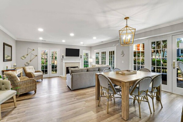 A spacious living room with a fireplace, a dining area with a wooden table and chairs, and a large window with a view of greenery.