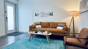 Un salon avec un canapé marron, une table basse, un tapis bleu, un lampadaire et une porte donnant sur une vue sur la montagne.