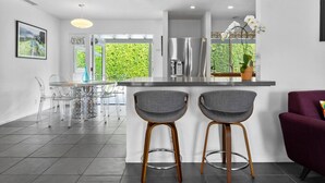 A modern kitchen with a central island, bar stools, a dining area with a glass table, and a view of greenery outside. - Stylish PS Home w/heated pool/spa & mountain views (Palm Springs)