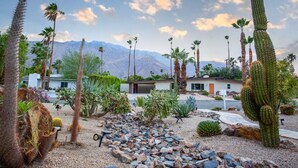 A desert garden with cacti and palm trees, a modern house, and mountains in the background. - Stylish PS Home w/heated pool/spa & mountain views (Palm Springs)