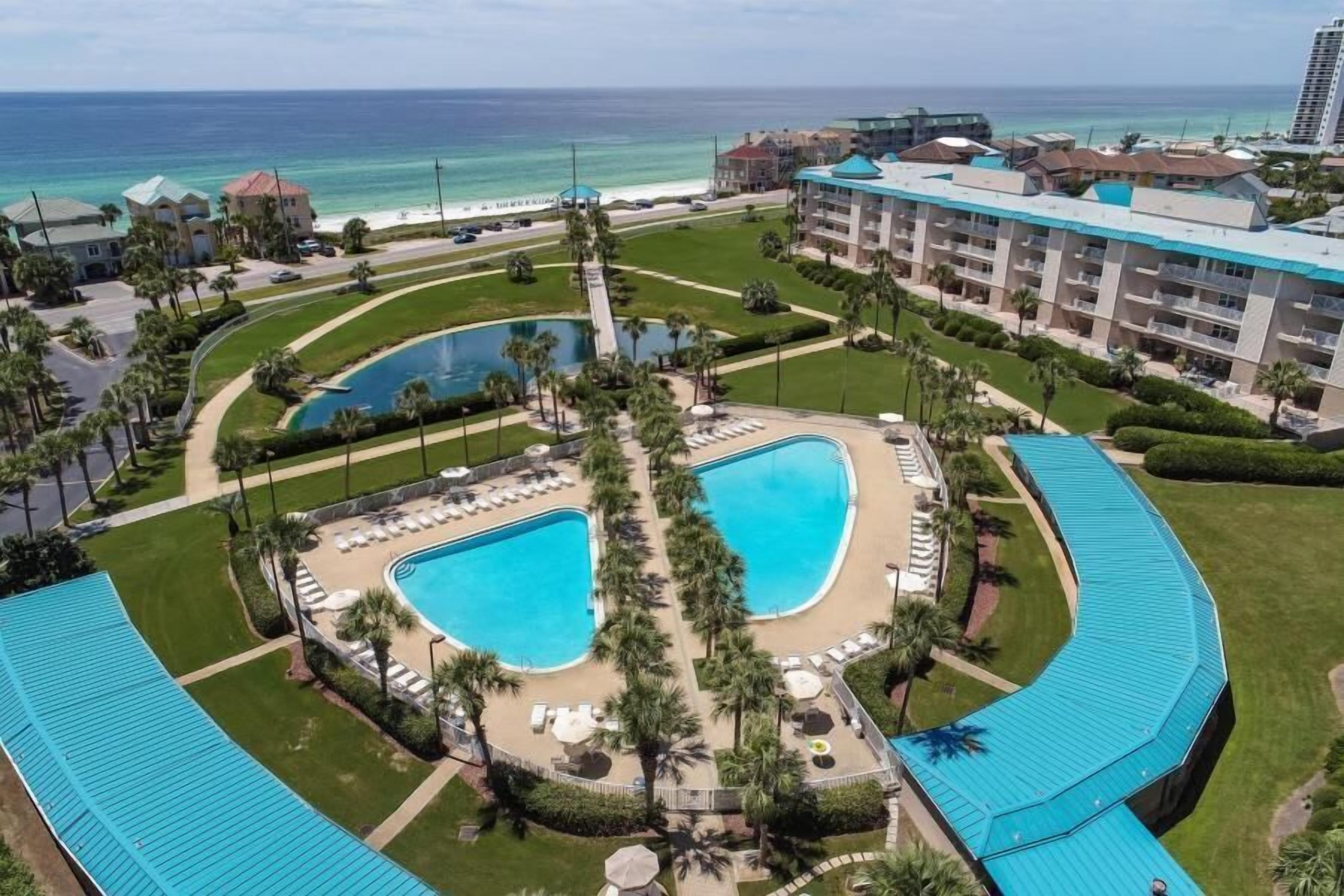 Un resort con dos piscinas grandes, vista a la playa y un edificio de varios pisos con techo azul.