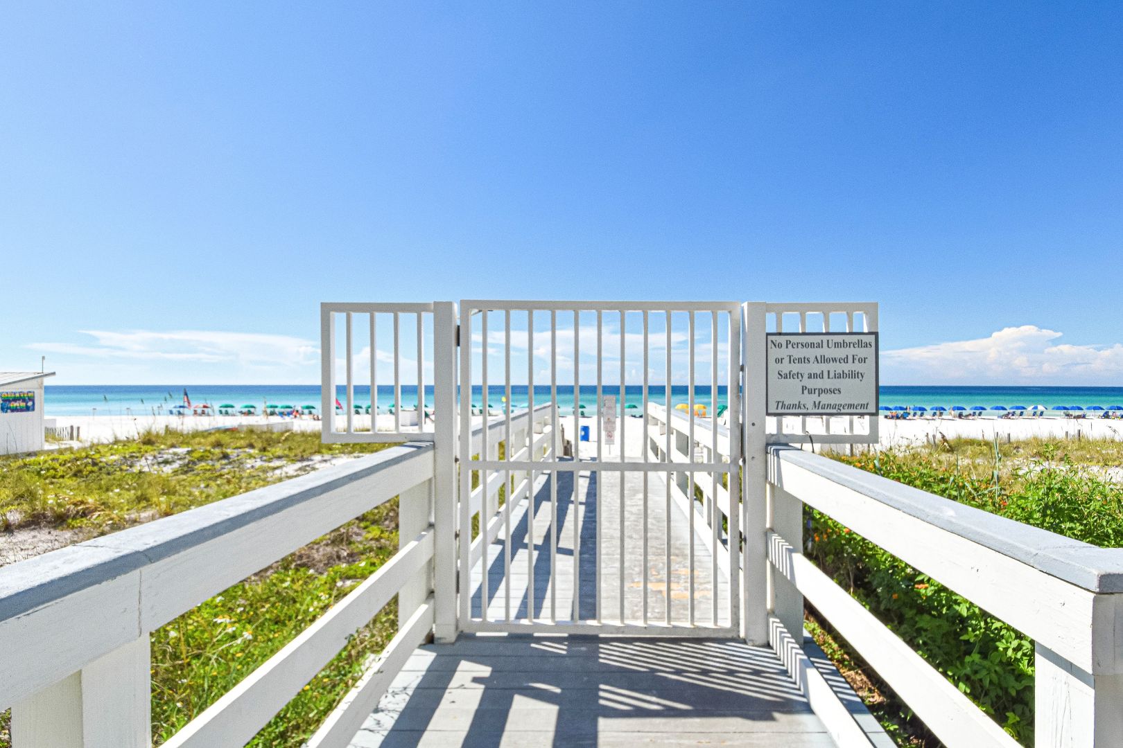 L'accesso alla spiaggia con un cancello bianco, un cielo azzurro limpido e un cartello che vieta l'uso di ombrelloni o tende personali.