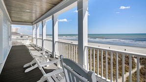 A covered porch with white railings and wooden deck chairs facing a beach.