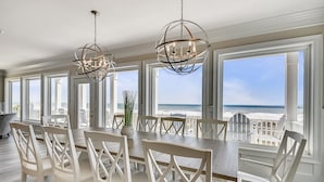 A spacious dining area with a large table and white chairs, large windows with a view of the ocean, and two chandeliers.