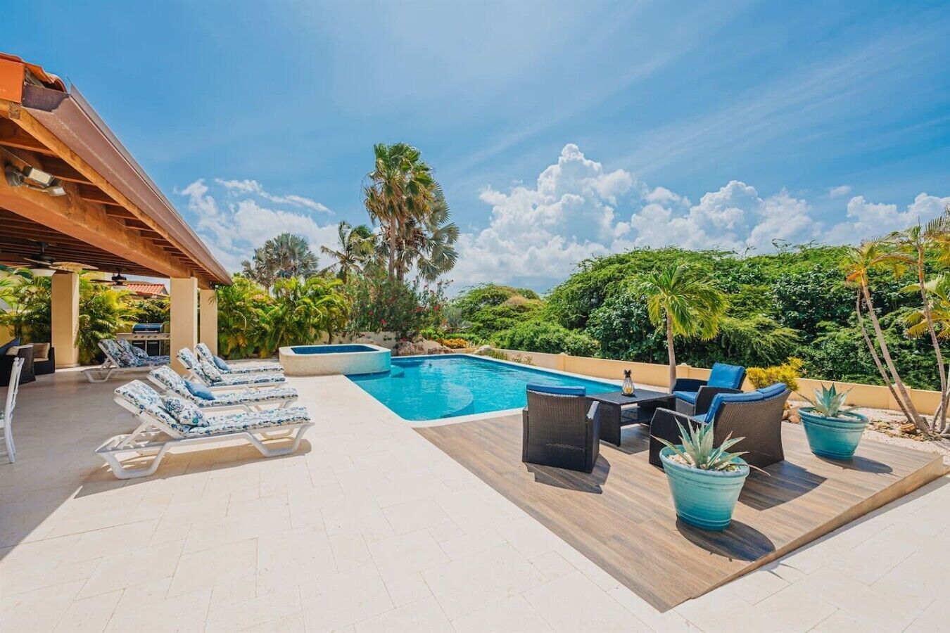 A pool area with lounge chairs and a table, surrounded by greenery and a clear sky.
