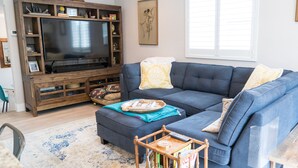 Ein Wohnzimmer mit einem großen Flachbildfernseher, einem blauen Sofa, einer Holztrennelemente und einem Fenster mit Jalousien.
