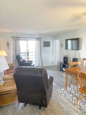 A living room with a leather sofa, a dining table with chairs, a TV mounted on the wall, and a window with curtains. - Two bedroom getaway near Tybee Beach & Lighthouse (Tybee Island)