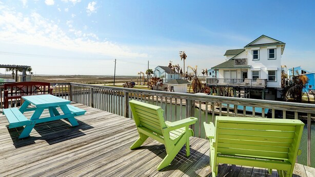 A wooden deck with picnic tables and chairs, overlooking a beach and a row of houses. - Jamaica Beach Waterfront Home! w/ Paddleboards, Kayak, Pedal Boat, Fishing Gears (Jamaica Beach)